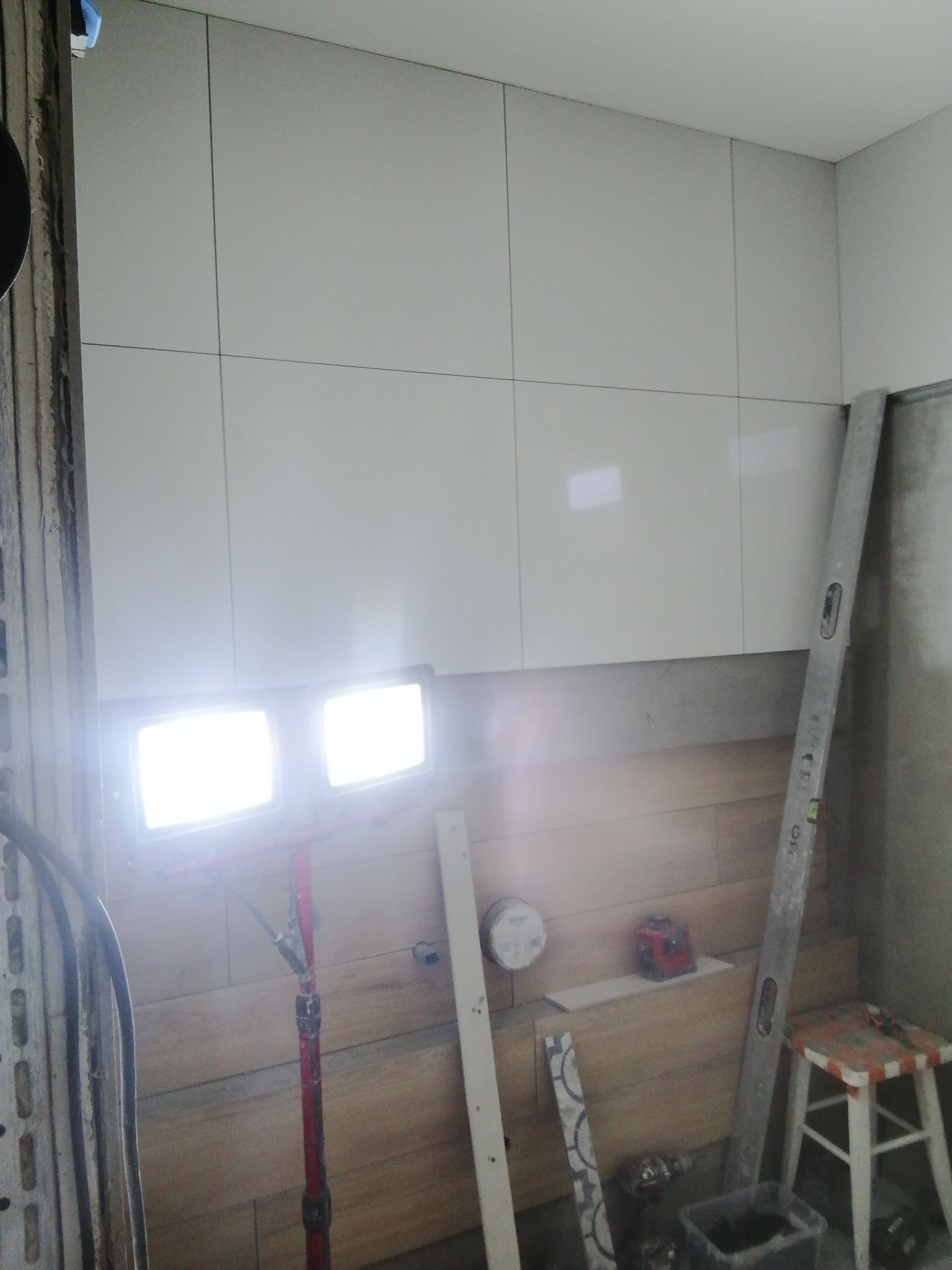 Montaż białych, kwadratowych płytek na ścianie, widoczna poziomica, reflektory oświetlające miejsce pracy, fragment ściany wyłożony płytkami imitującymi drewno, czerwony laser krzyżowy i taboret...