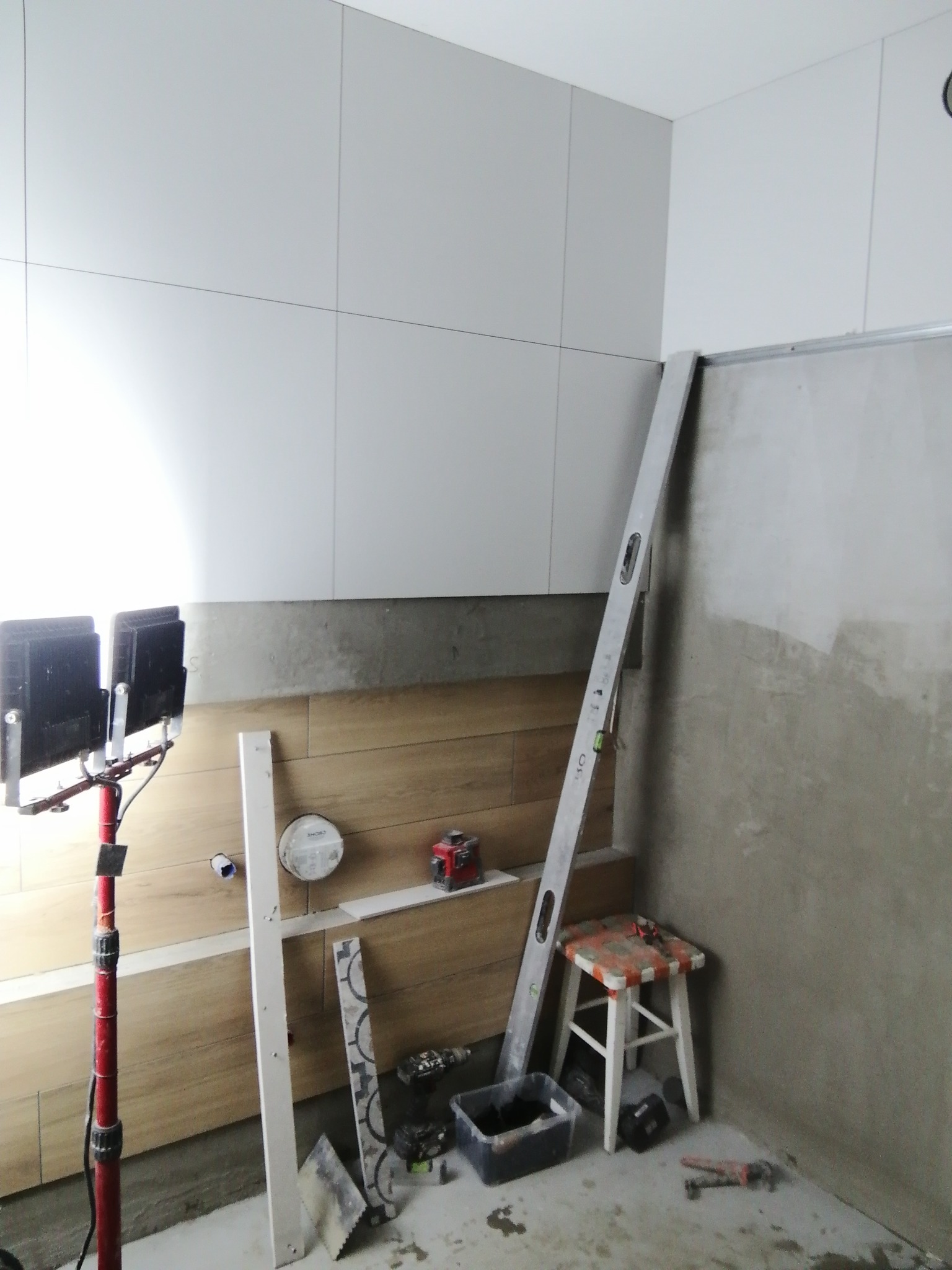 Łazienka w trakcie układania płytek: białe płytki na górze, drewnopodobne na dole, poziomica oparta o ścianę, lampa budowlana oświetla obszar roboczy, widoczne narzędzia glazurnicze, mały stołek.