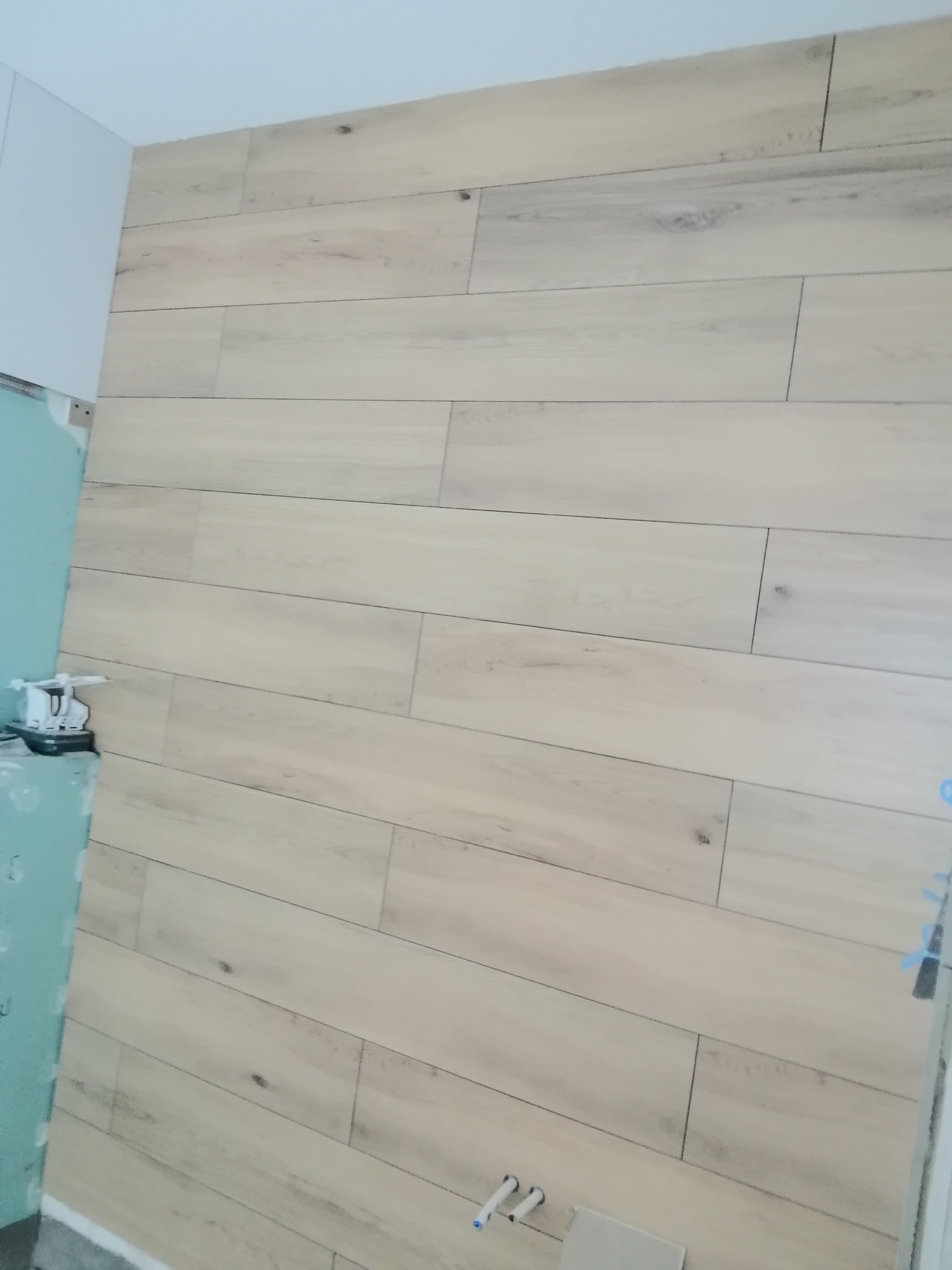Ściana wyłożona jasnymi, drewnopodobnymi płytkami, ułożonymi poziomo, z widocznymi fragmentami zielonej płyty gipsowo-kartonowej i instalacji hydraulicznej w dolnym rogu.