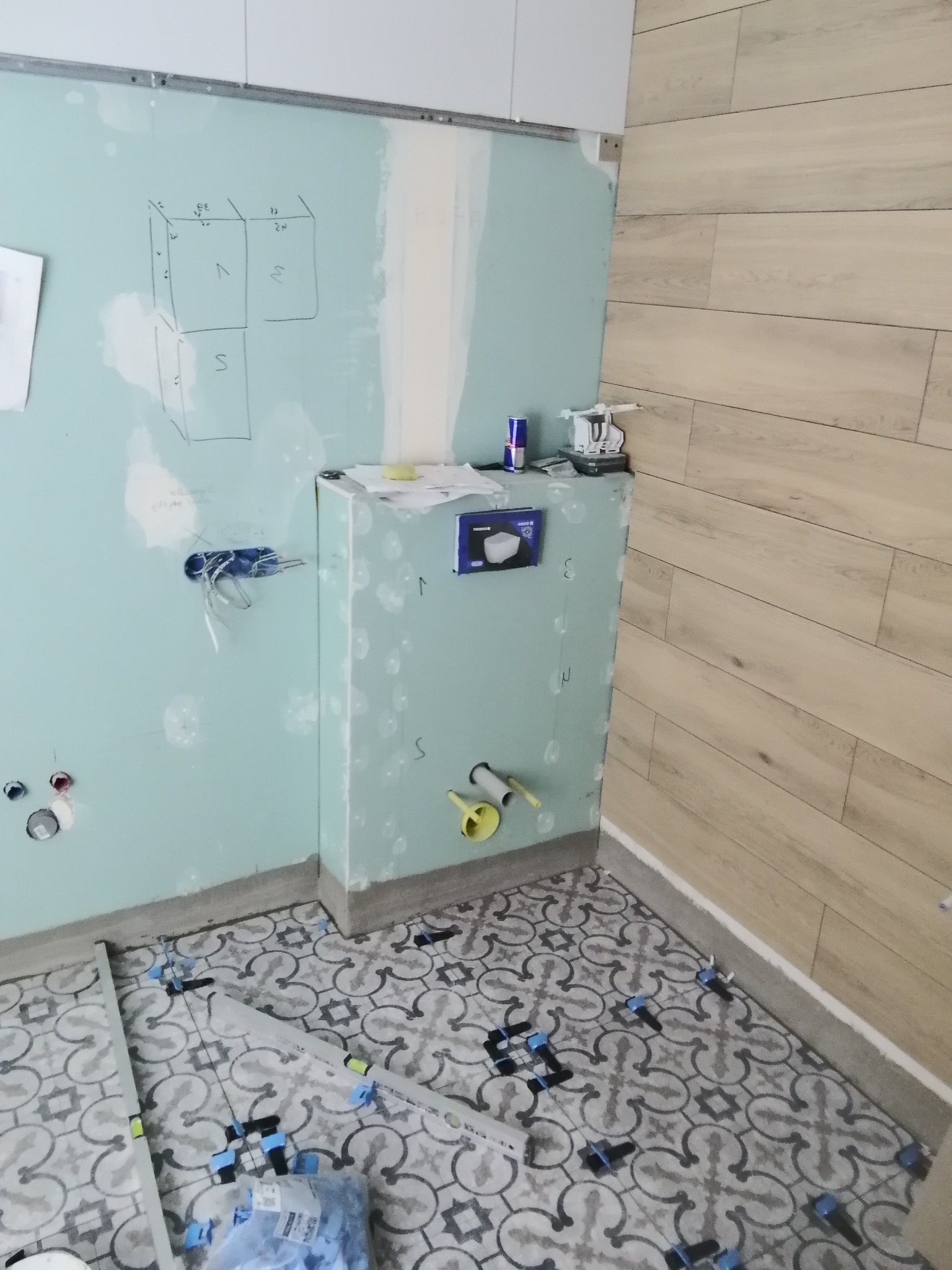Łazienka w trakcie remontu: położone płytki podłogowe z wzorem, ściany częściowo wykończone płytami gipsowo-kartonowymi z zaznaczonymi punktami i schematem na ścianie, widoczne rury i instalacje...