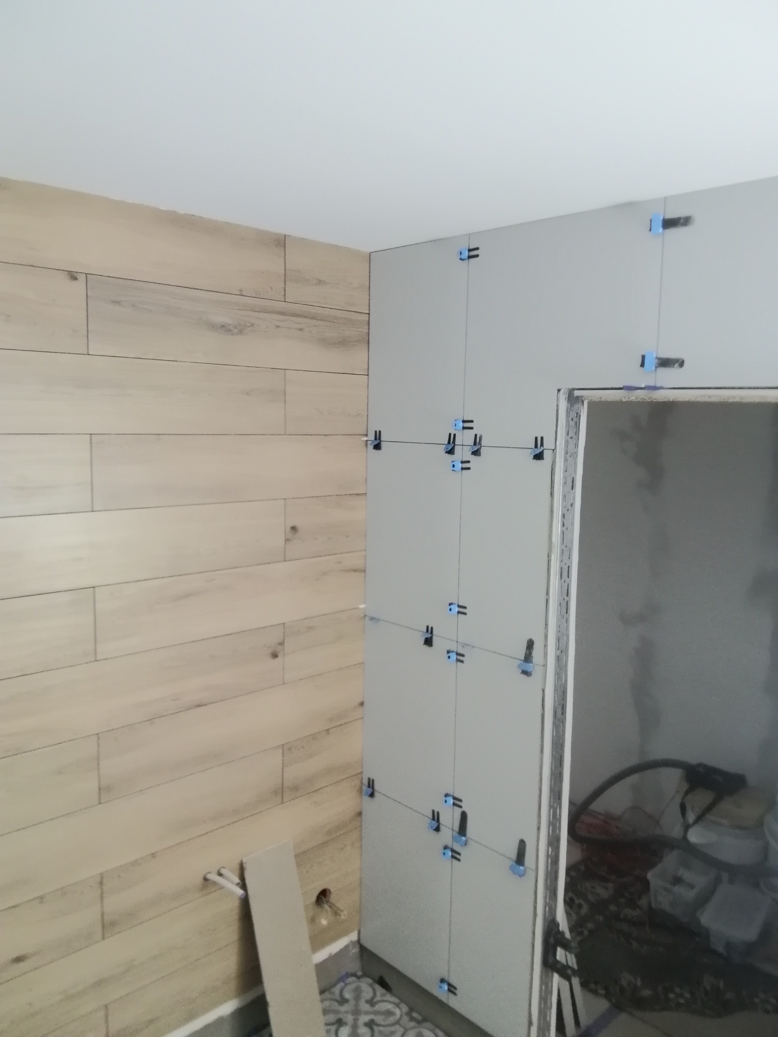 Fragment ściany z płytkami imitującymi drewno oraz fragment ściany przygotowanej pod płytki z użyciem niebieskich krzyżyków dystansowych, widoczne rury i fragment podłogi z wzorem.