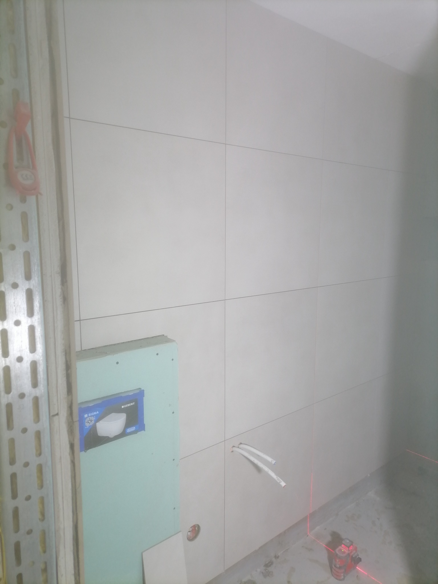 Ściana łazienki w trakcie układania jasnych płytek, widoczny laser krzyżowy, przyłącza do instalacji sanitarnej oraz zabudowa stelaża podtynkowego Geberit Sigma.