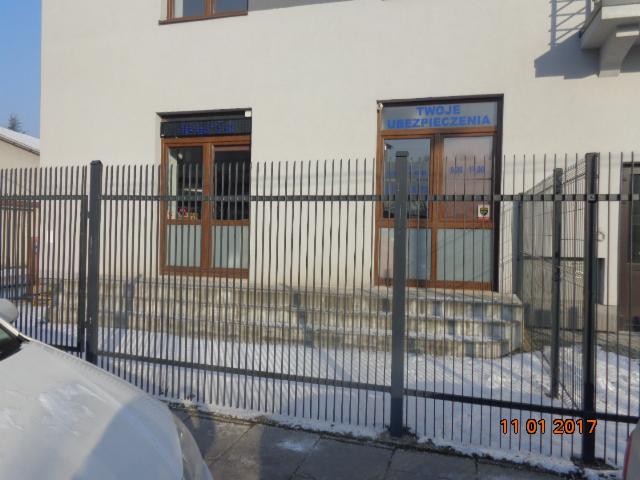 Fasada budynku z dwoma wejściami, na oknach napisy 'UBEZPIECZENIA' i 'TWOJE UBEZPIECZENIA', widoczne przez metalowe ogrodzenie w zimowej scenerii.