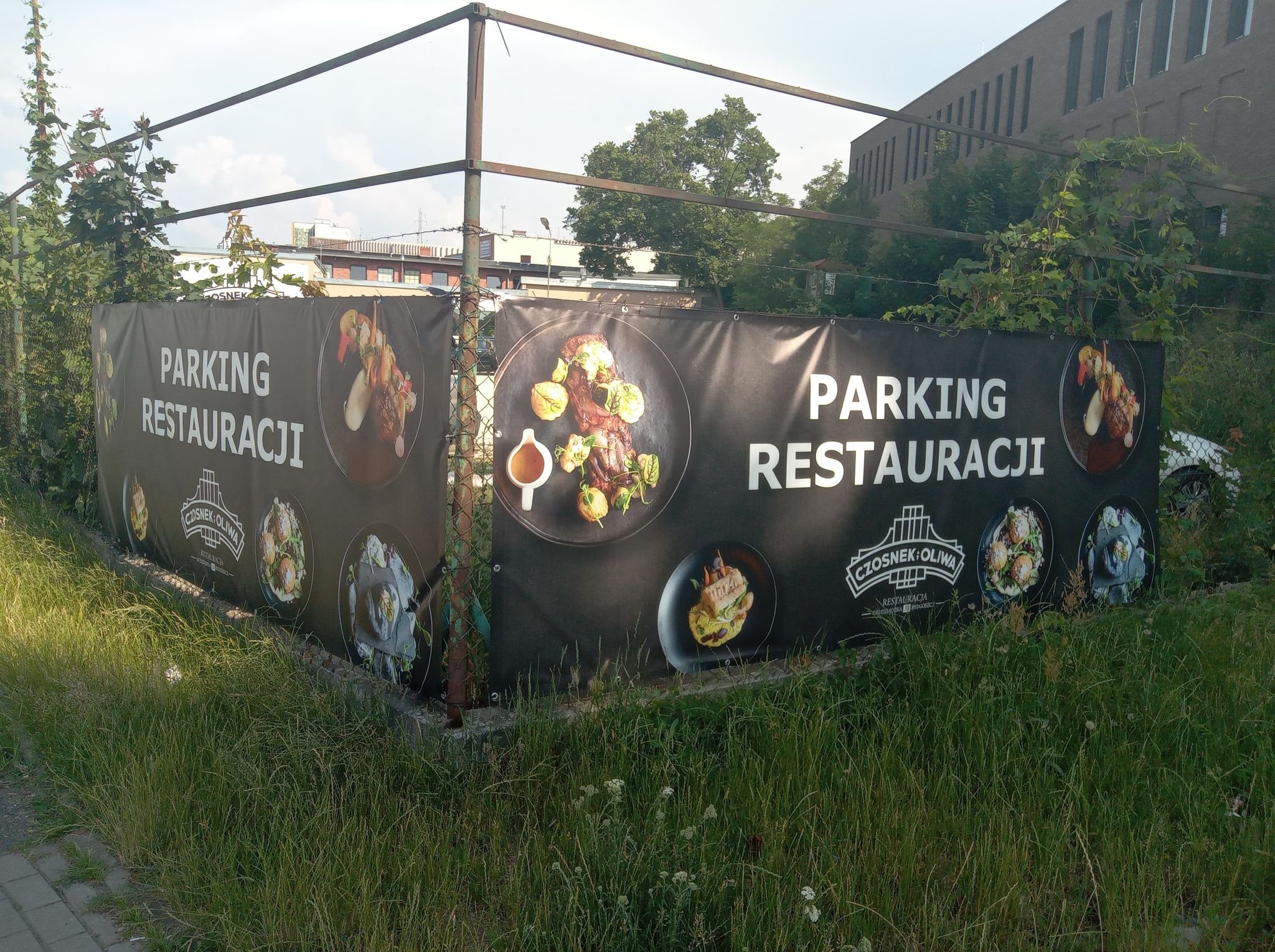 Czarny baner reklamowy z napisem 'Parking Restauracji' i zdjęciami dań, umieszczony na metalowej konstrukcji przy parkingu, zarośnięty trawą.