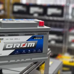 Szary akumulator samochodowy marki GRØM z czerwonym i czarnym korkiem na tle półek sklepowych z innymi akumulatorami.