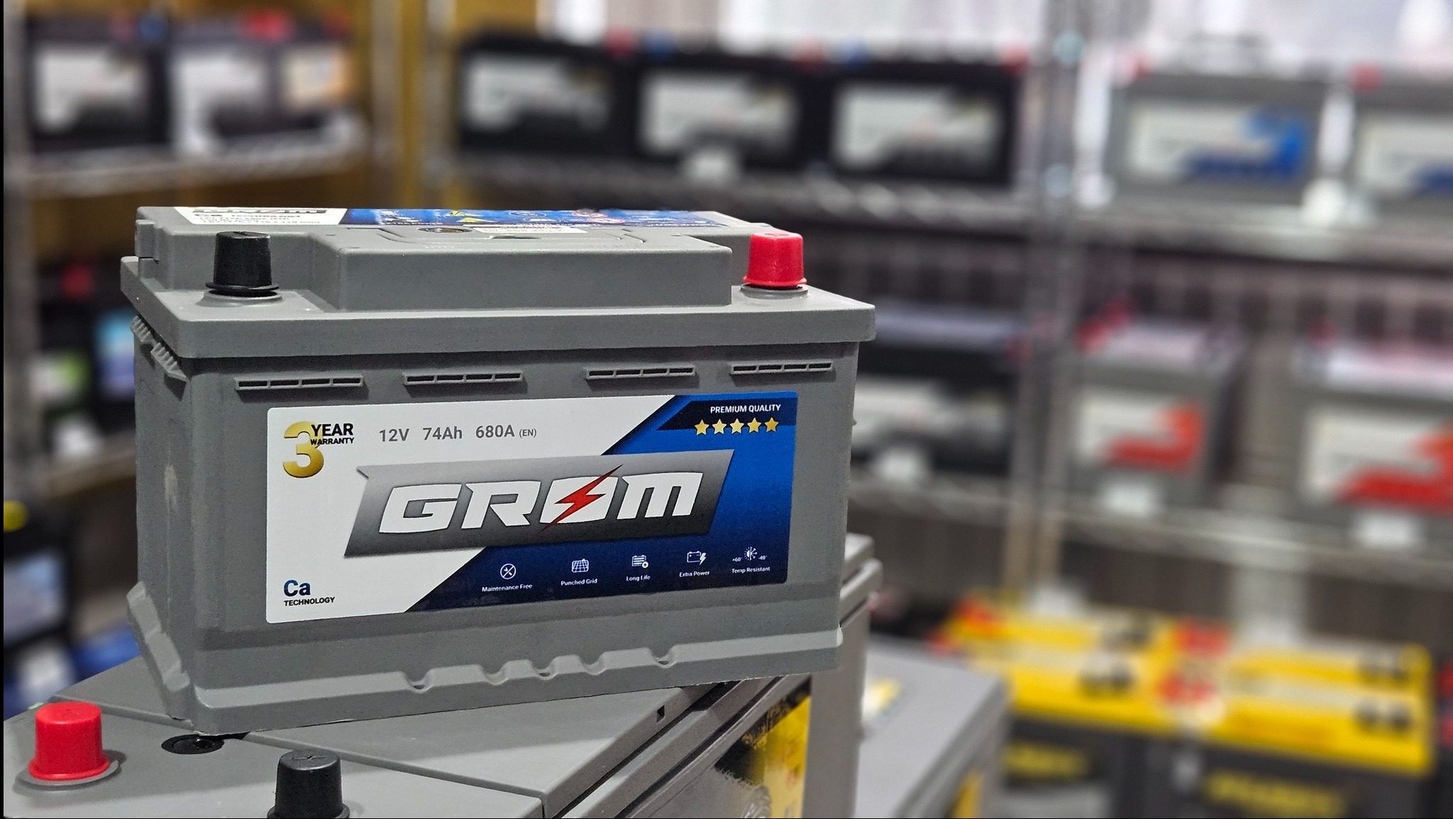 Szary akumulator samochodowy marki GRØM z czerwonym i czarnym korkiem na tle półek sklepowych z innymi akumulatorami.