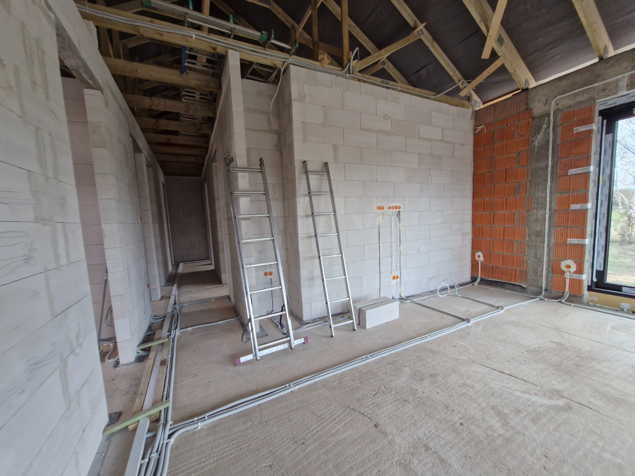 Wnętrze w trakcie budowy z widocznymi przewodami w peszlach na podłodze i wyprowadzonymi punktami elektrycznymi w ścianach. Dwie drabiny opierają się o ścianę.