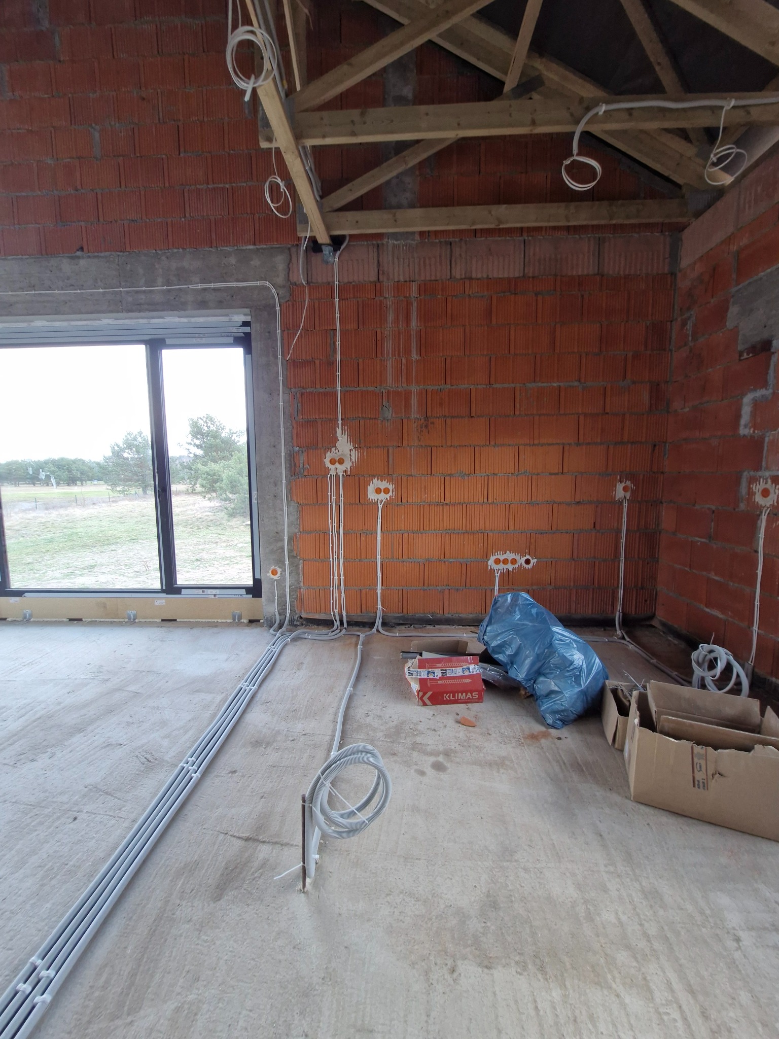 Surowy stan: wnętrze z ceglanymi ścianami w trakcie prac, widoczne kable i puszki elektryczne, okno z widokiem na zewnątrz, podłoga betonowa, elementy drewniane pod dachem.