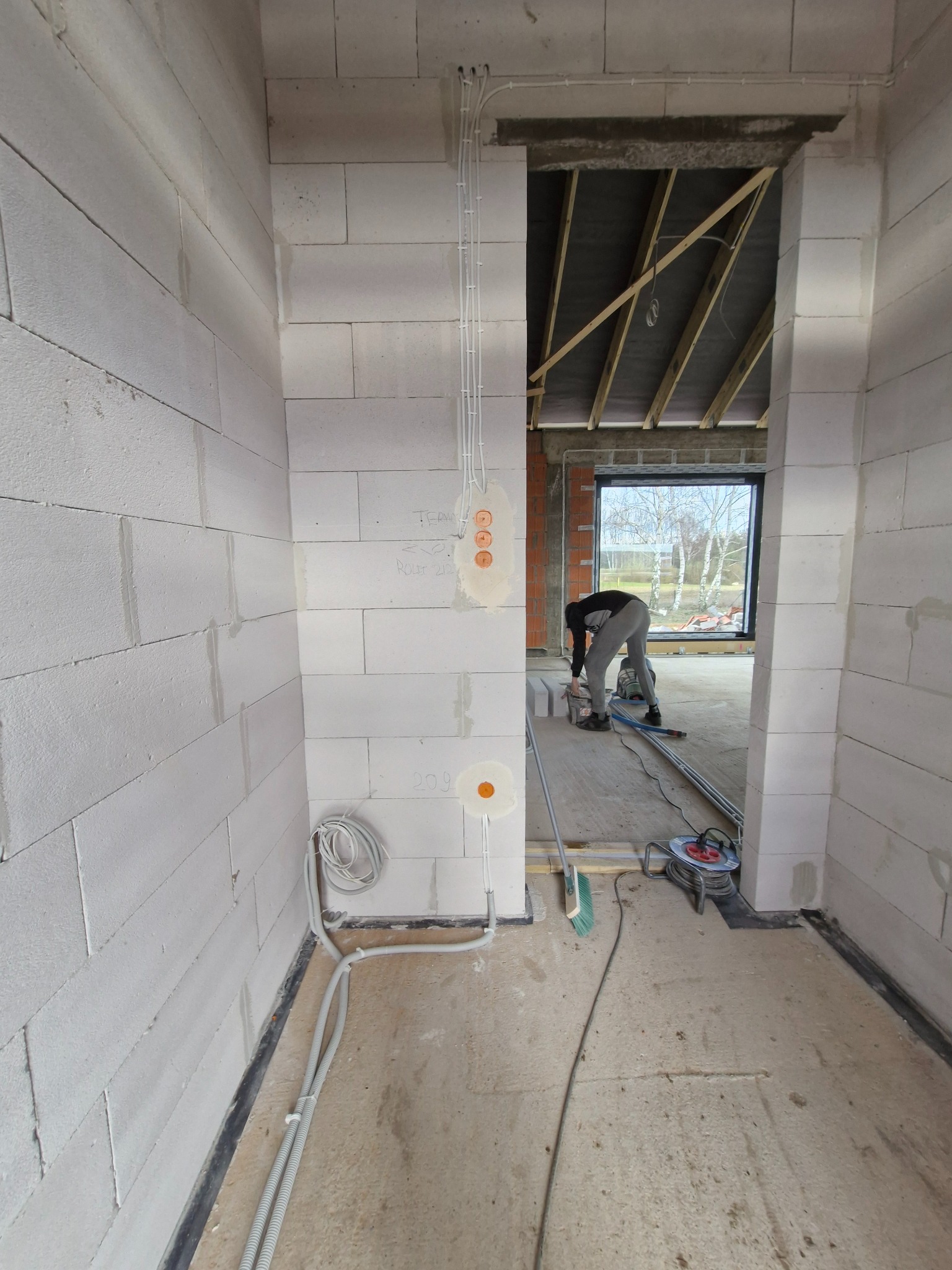 Wnętrze w trakcie budowy z widocznymi przewodami w ścianach i podłodze, przygotowane pod gniazdka. W tle osoba przy pracy i okno z widokiem na zewnątrz.