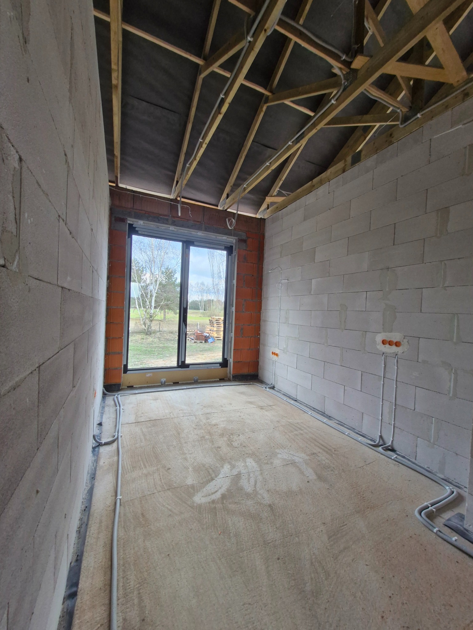 Wnętrze w stanie surowym z widoczną instalacją elektryczną w peszlach, gniazda w ścianie z betonu komórkowego, okno z widokiem na zewnątrz, drewniana konstrukcja dachu.