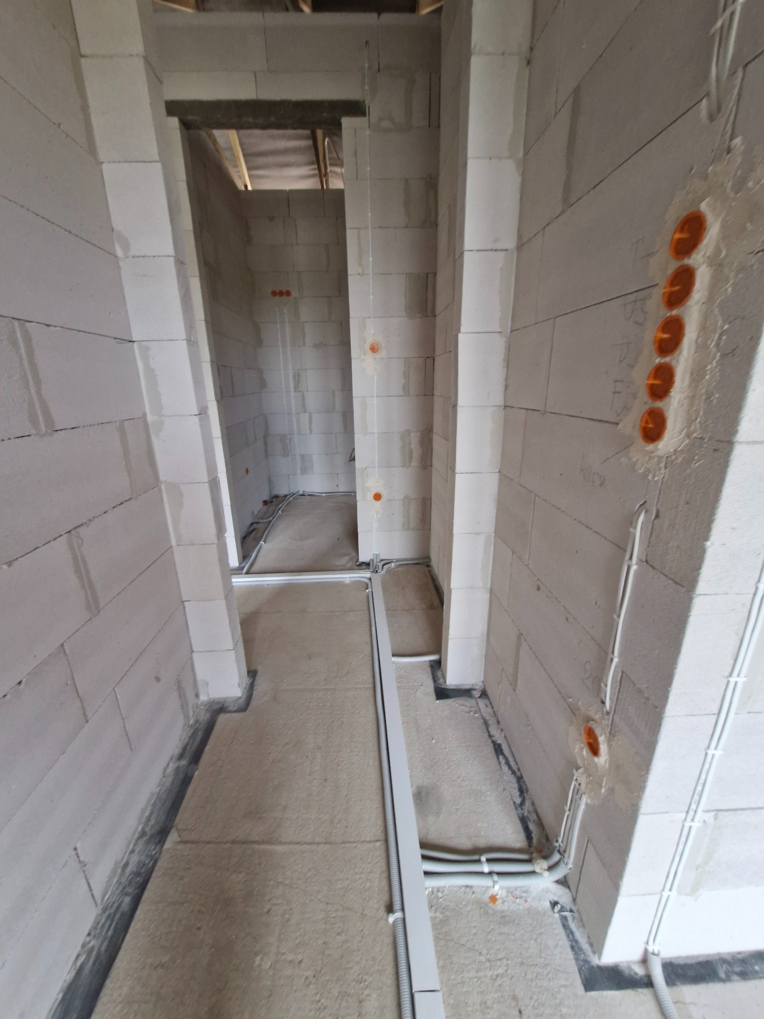 Wnętrze w trakcie budowy z widocznymi przewodami elektrycznymi ułożonymi wzdłuż ścian i podłogi, oraz pomarańczowymi puszkami instalacyjnymi w ścianie.