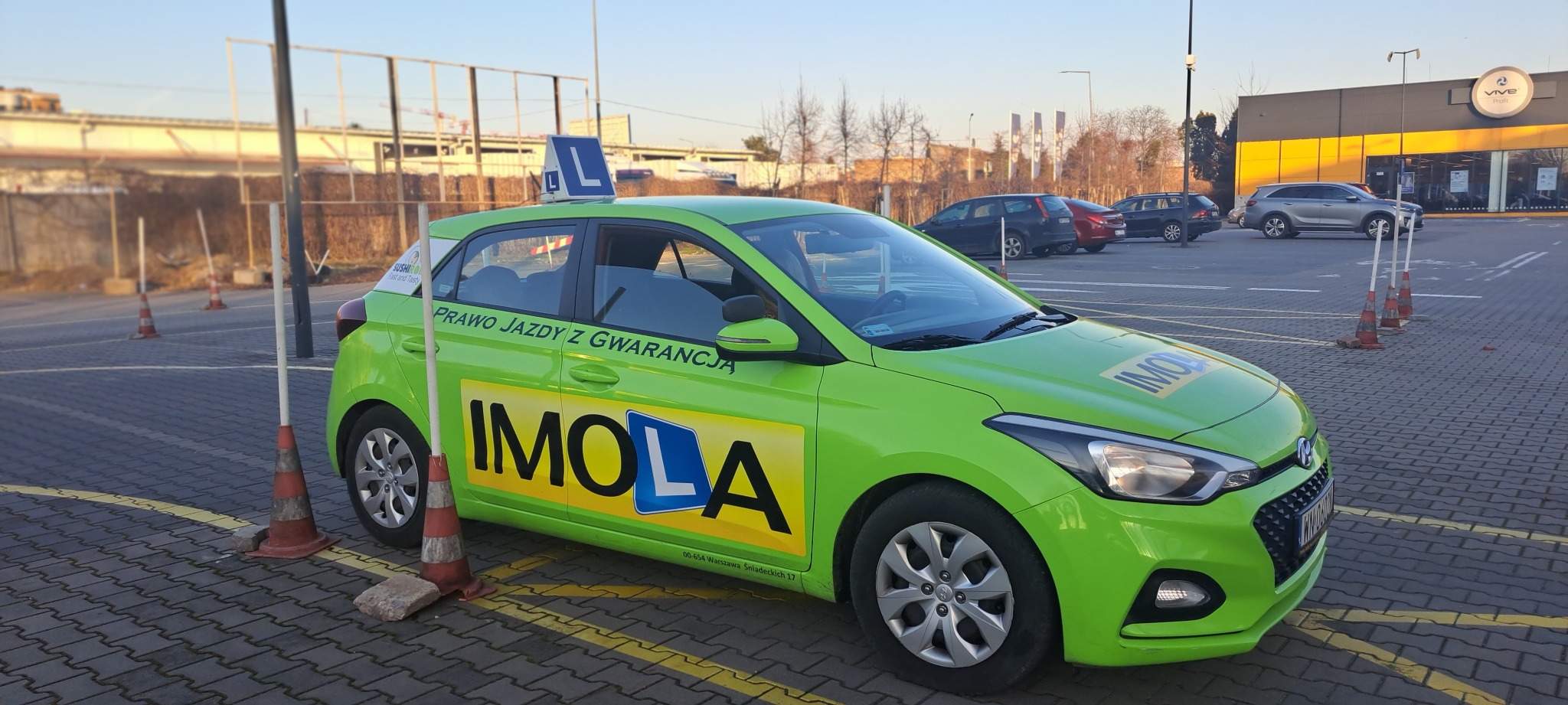 Jaskrawo zielony samochód szkoły jazdy IMOLA z niebieską literą 'L' na dachu, zaparkowany na parkingu z pomarańczowymi pachołkami i budynkiem Vive Profit w tle.