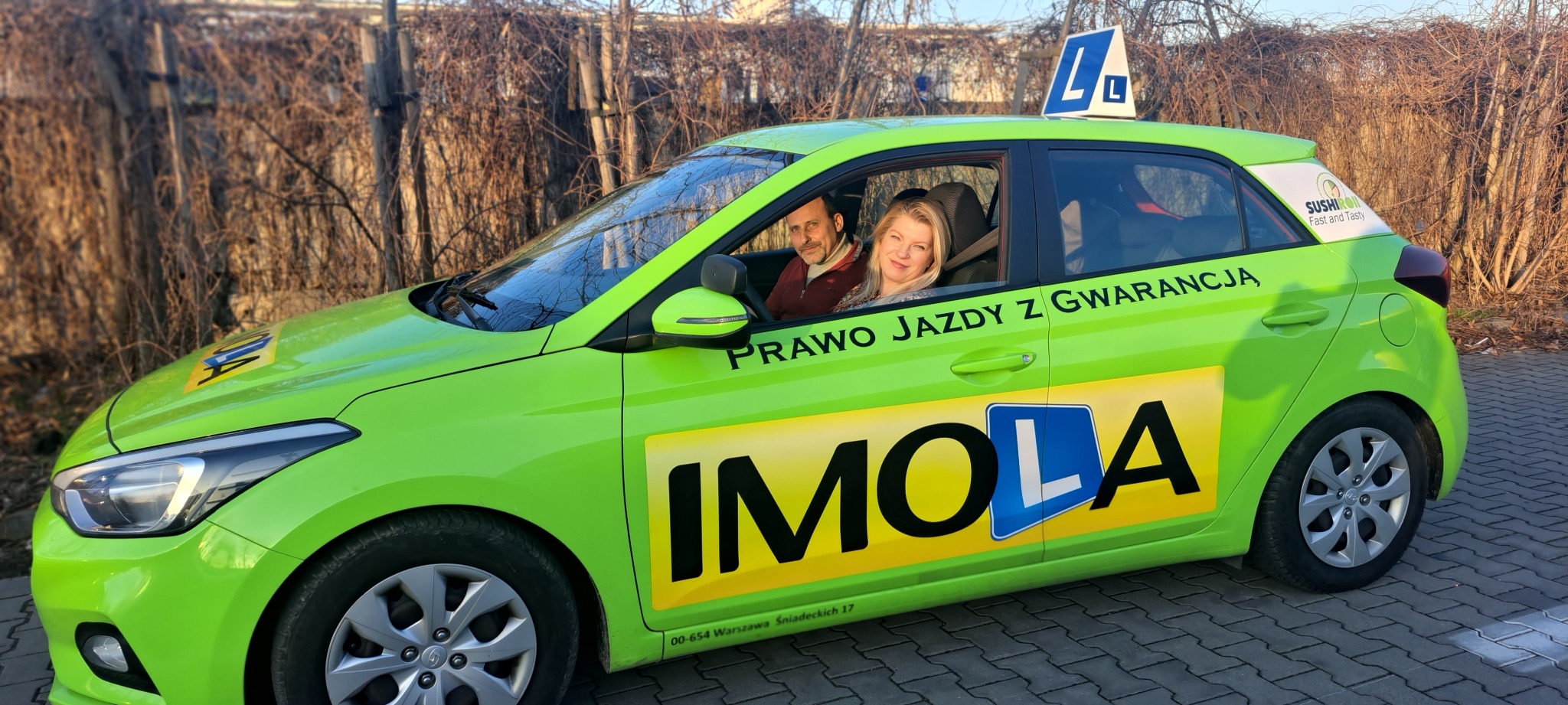 Jaskrawozielony samochód z oznaczeniami 'L' na dachu i na karoserii, z napisem 'Prawo jazdy z gwarancją' oraz nazwą 'IMOLA', instruktor i kursantka w środku, na tle drzew bez liści.