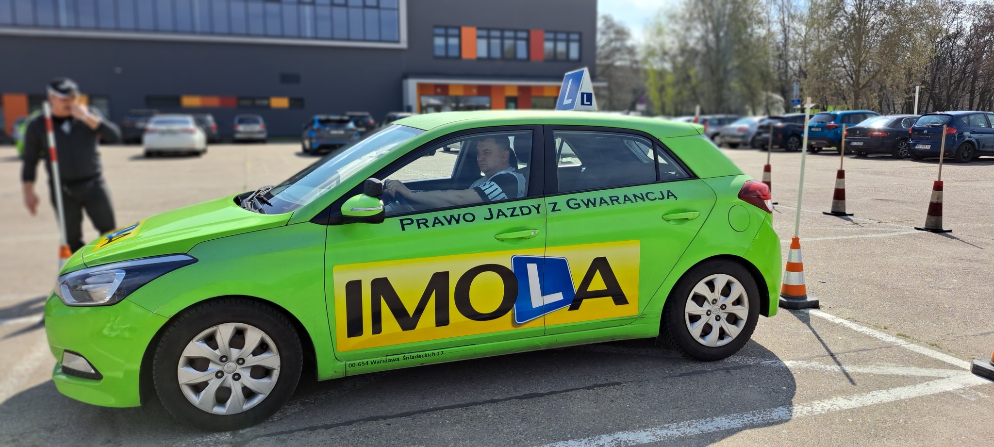 Zielony samochód z oznaczeniem nauki jazdy 'L', zaparkowany na placu manewrowym z pachołkami, instruktor z tyczką w tle.