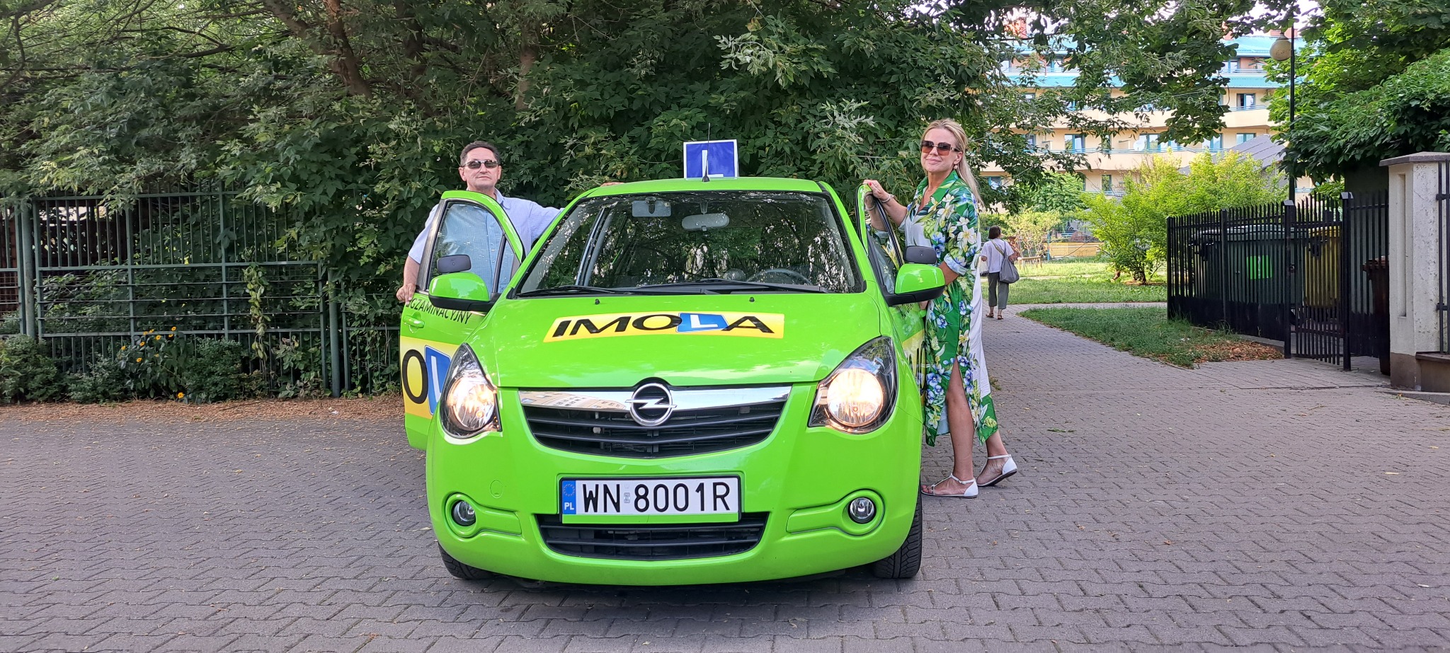 Jaskrawozielony samochód z oznaczeniami firmy IMOLA, służący do nauki jazdy, z instruktorem i kursantką obok, na tle zieleni i budynku mieszkalnego.