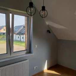 Pokój na poddaszu po remoncie: świeżo pomalowane szare ściany, drewniana podłoga, nowoczesne czarne lampy wiszące i okno z widokiem na osiedle domów.