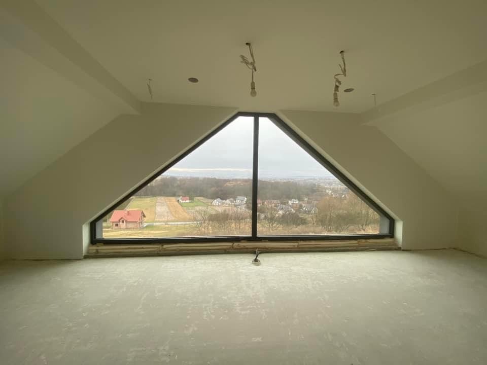 Poddasze w stanie deweloperskim z trójkątnym oknem, widok na krajobraz z zabudową jednorodzinną, surowe ściany i podłoga przygotowane do dalszych prac wykończeniowych, wiszące przewody oświetleniowe.