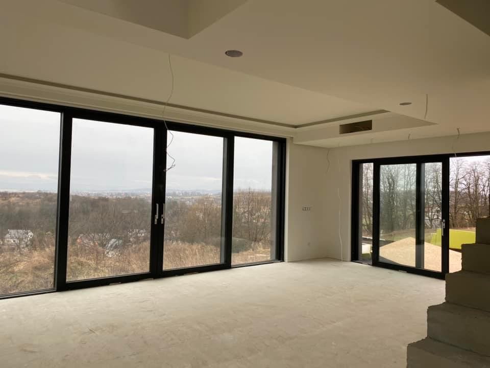 Wnętrze w stanie deweloperskim z dużymi przesuwnymi oknami z czarnymi ramami, widok na panoramę miasta w oddali, surowa podłoga, widoczne przewody elektryczne