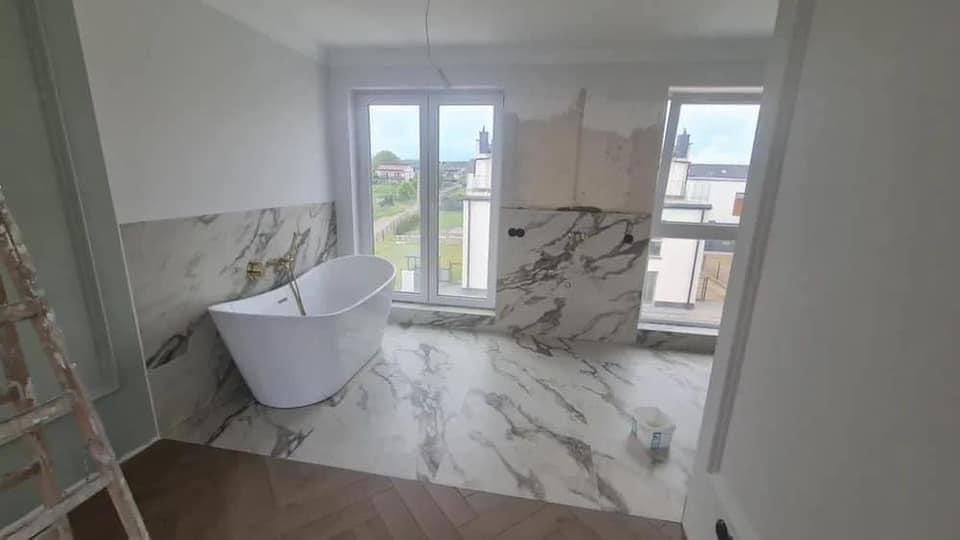 Wnętrze łazienki w trakcie wykańczania: biała wolnostojąca wanna, marmurowe płytki na podłodze i ścianach do połowy wysokości, widoczne niedokończone fragmenty ścian, drabina malarska z lewej...