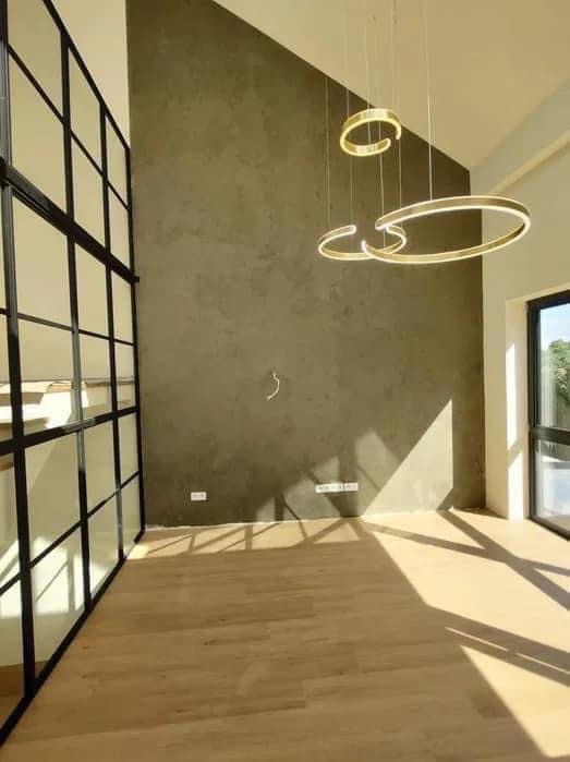 Nowoczesne wnętrze z drewnianą podłogą, ścianą w odcieniach szarości, przeszkloną ścianą działową z czarnymi profilami i wiszącymi lampami w kształcie złotych pierścieni.