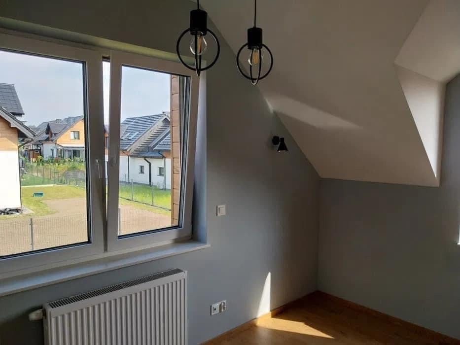 Pokój na poddaszu po remoncie: świeżo pomalowane szare ściany, drewniana podłoga, nowoczesne czarne lampy wiszące i okno z widokiem na osiedle domów.