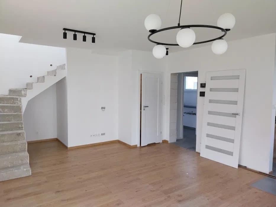 Wnętrze w trakcie prac wykończeniowych: surowe schody betonowe, świeżo położone panele podłogowe, białe ściany, nowoczesne lampy sufitowe i białe drzwi z przeszkleniami.