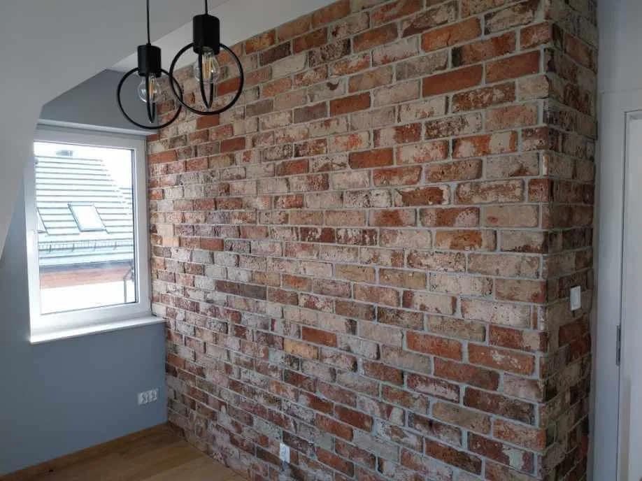 Ściana z cegły dekoracyjnej w pokoju, oświetlona przez wiszące lampy z żarówkami Edisona, widok z okna na dach sąsiedniego budynku.