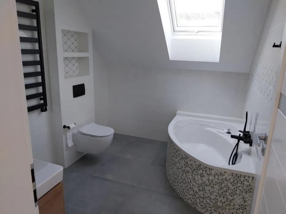 Nowoczesna łazienka na poddaszu z oknem dachowym, szare płytki podłogowe, wanna obudowana mozaiką, wisząca toaleta i czarny grzejnik drabinkowy.