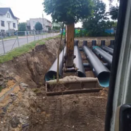 Żółta koparka umieszcza rury drenażowe z zielonym wnętrzem w wykopie, widok z kabiny, w tle drzewo i budynki mieszkalne.