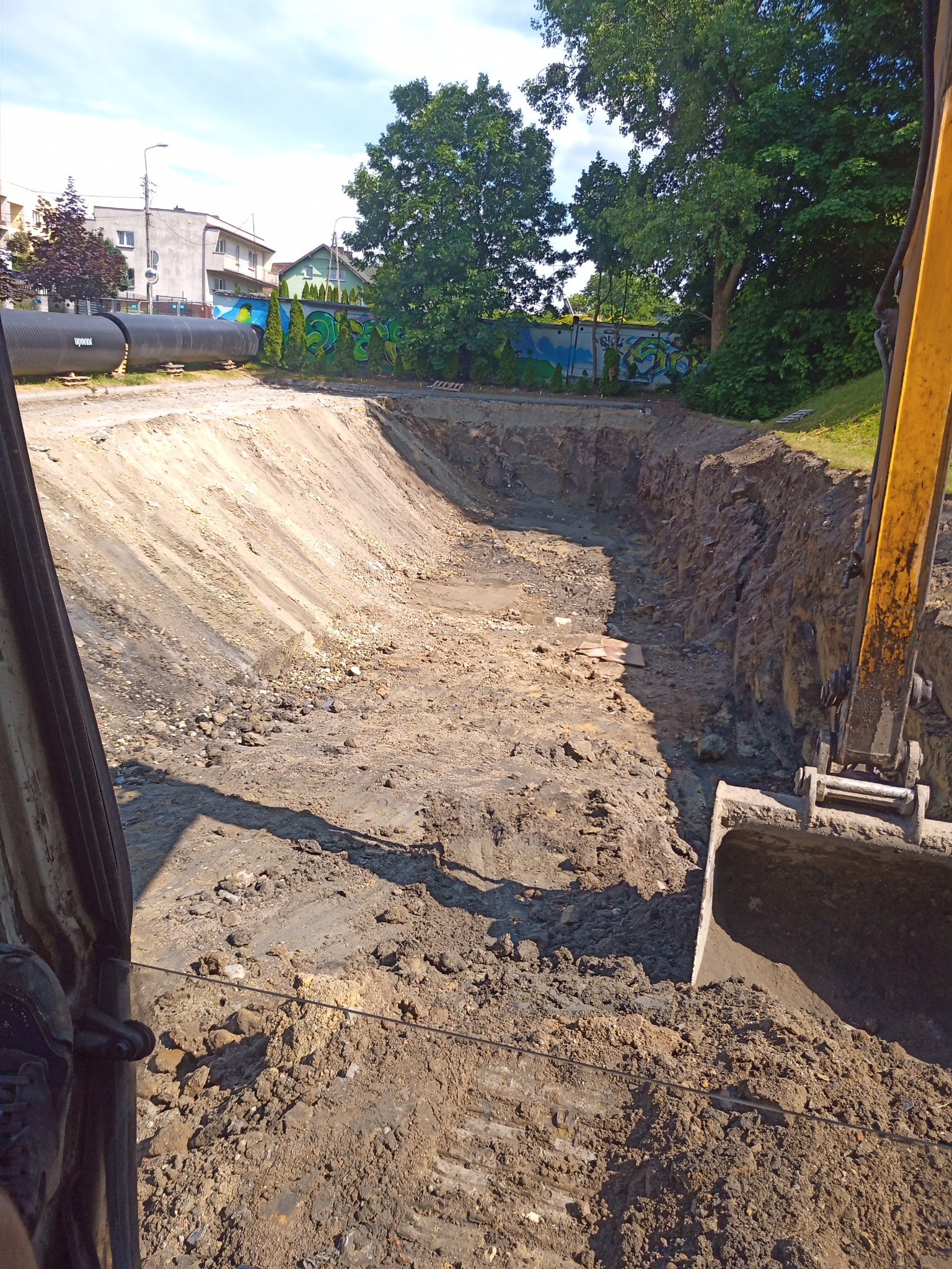 Głęboki wykop w ziemi z widocznymi śladami pracy koparki, obok stoi maszyna, w tle widoczne budynki mieszkalne, rury i zielona ściana z graffiti.