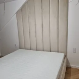 Beżowe, tapicerowane wezgłowie łóżka z pionowymi panelami w pokoju ze skośnym sufitem, widoczne gniazdko elektryczne na ścianie.