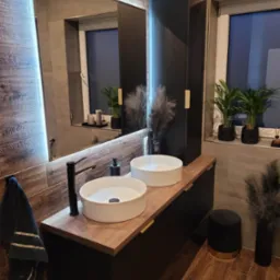 Nowoczesna łazienka z dwoma okrągłymi umywalkami nablatowymi, czarną armaturą i szafką z drewnianym blatem, uzupełniona oświetlonym lustrem i elementami dekoracyjnymi, takimi jak rośliny doniczkowe...