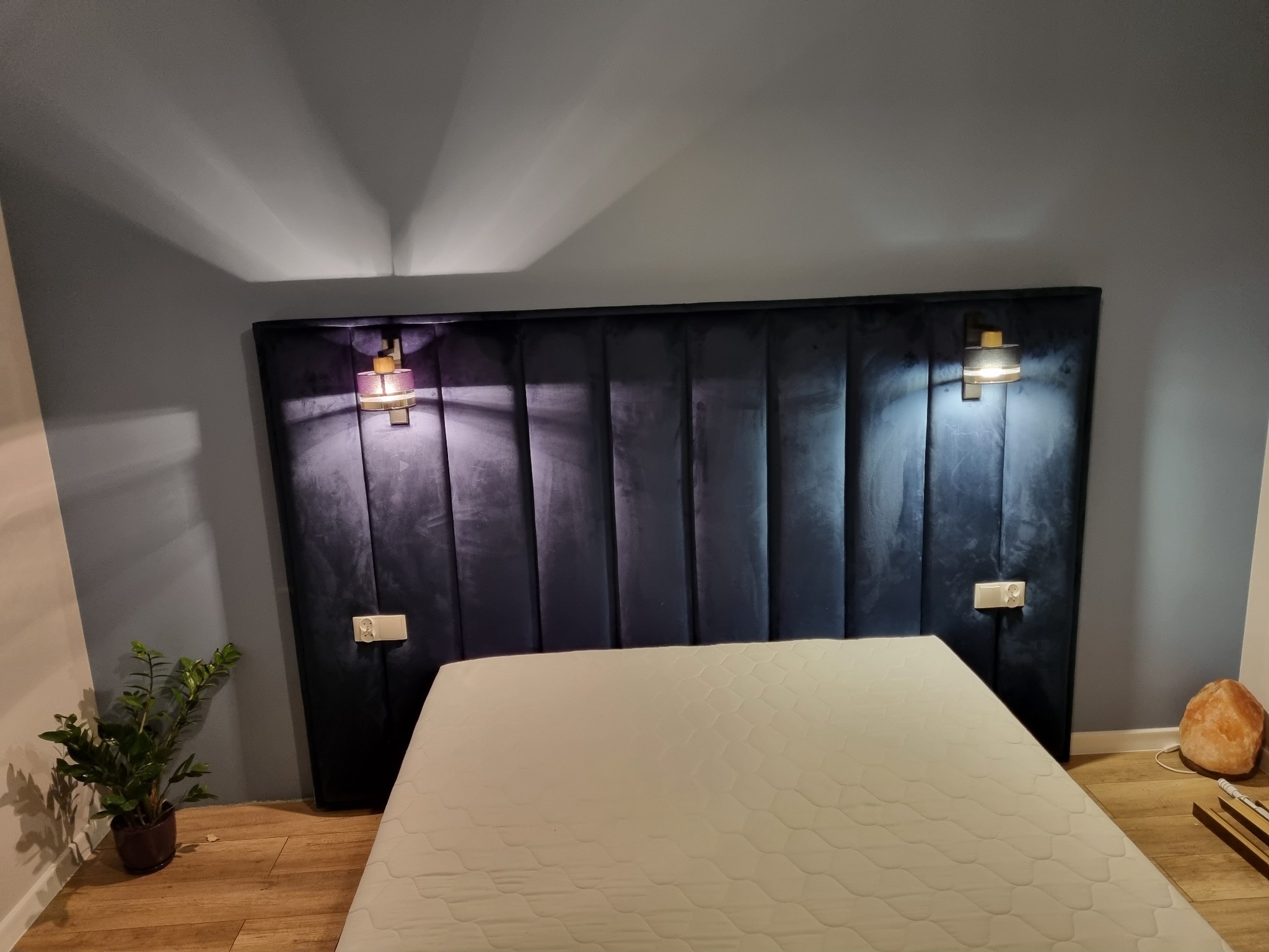 Tapicerowany zagłówek w kolorze granatowym z pionowymi panelami, oświetlony kinkietami z ciepłym światłem, ustawiony za materacem na podłodze, z widoczną rośliną doniczkową i lampą solną.