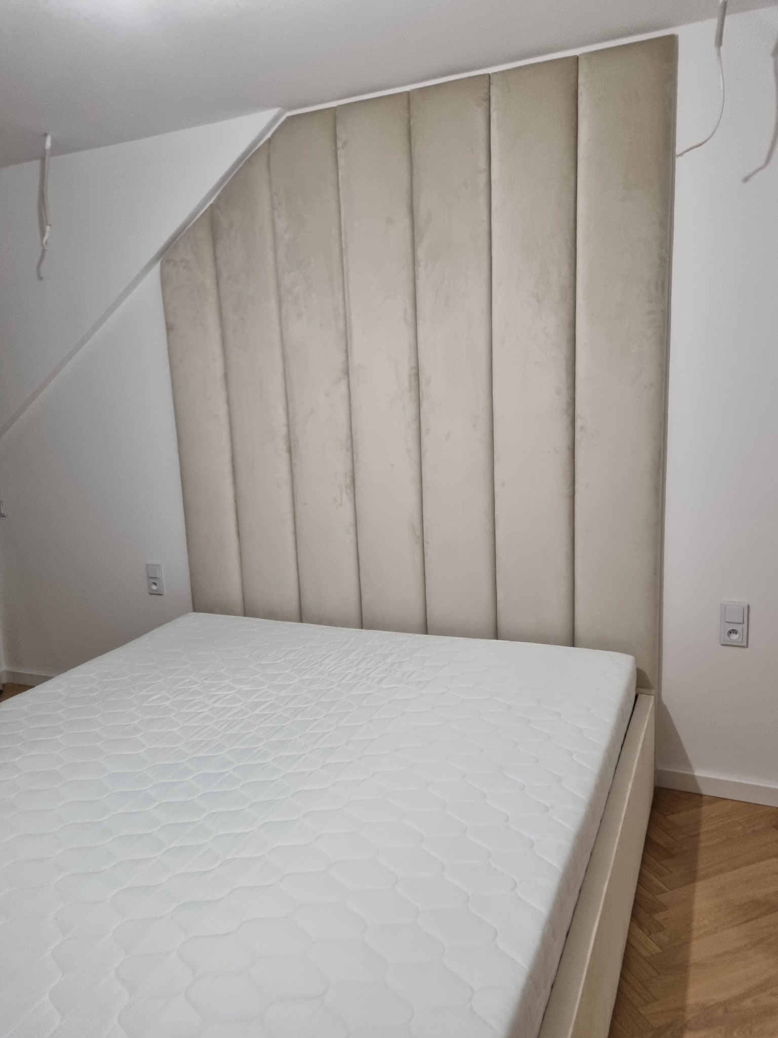 Beżowe, tapicerowane wezgłowie łóżka z pionowymi panelami w pokoju ze skośnym sufitem, widoczne gniazdko elektryczne na ścianie.