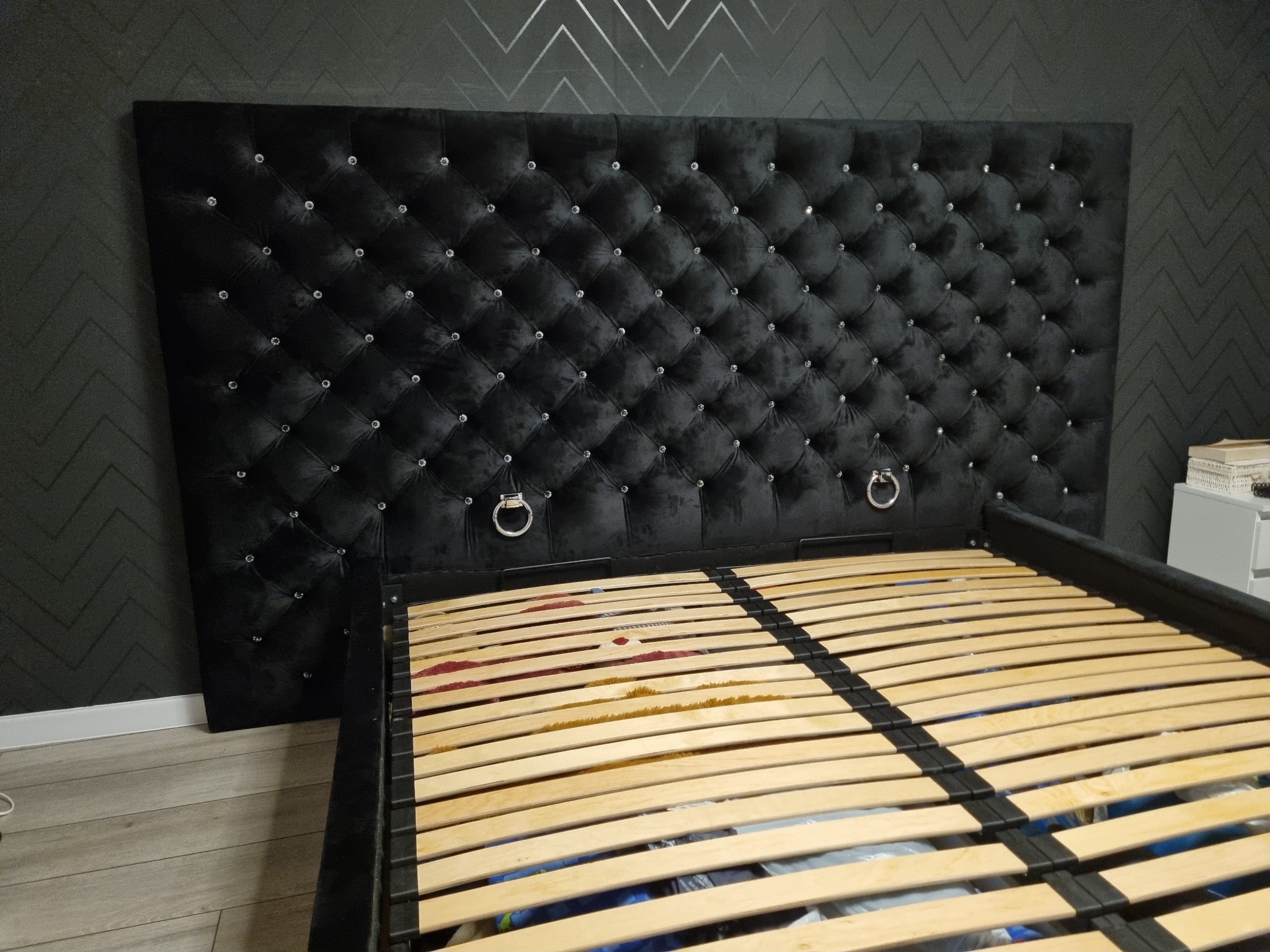 Czarne, pikowane wezgłowie łóżka z kryształkami i metalowymi uchwytami, widoczny stelaż z drewnianych listew, tło stanowi ciemna ściana w geometryczny wzór.