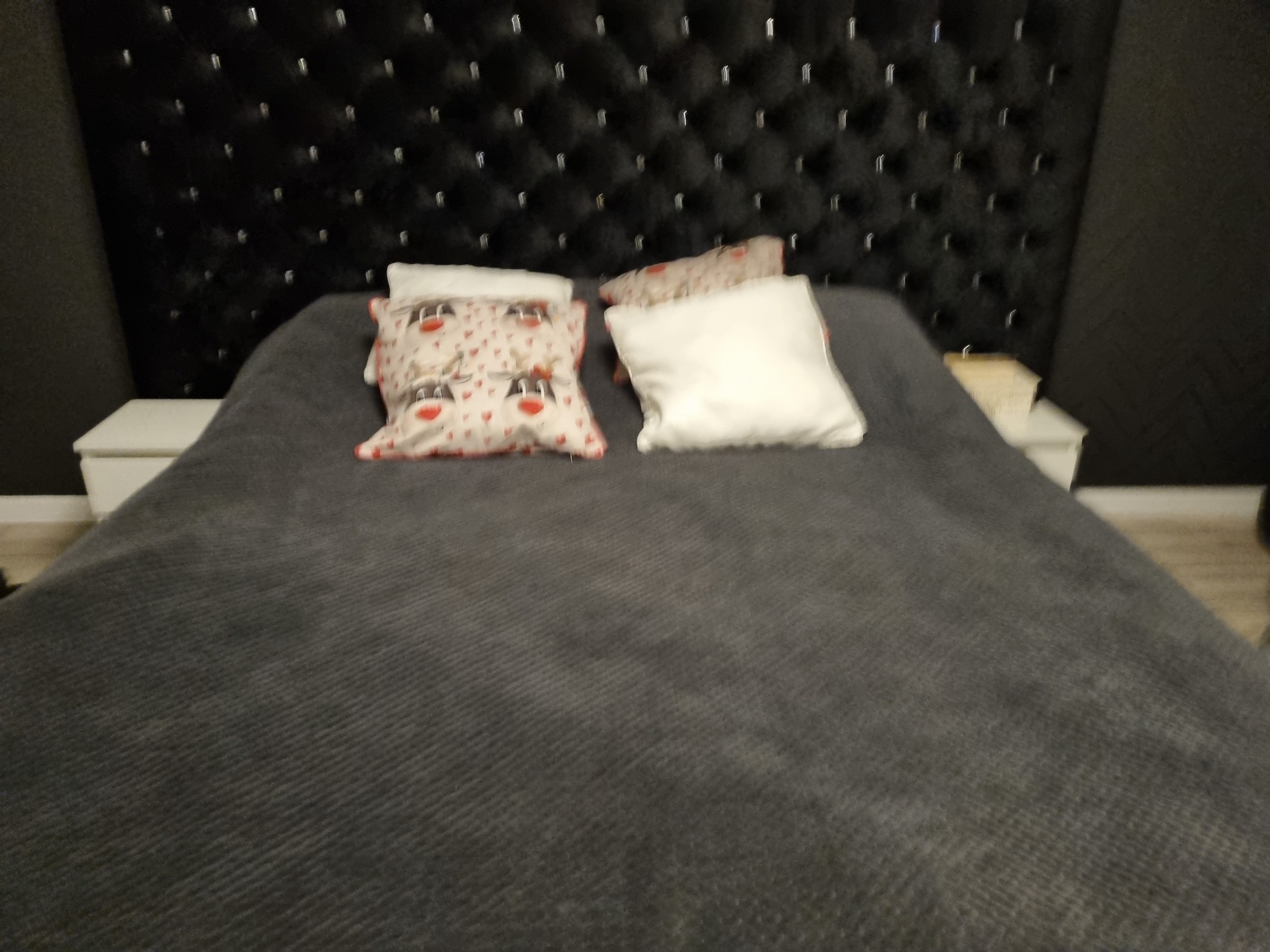 Łóżko z ciemnoszarą narzutą, poduszki z motywem reniferów i pikowane, czarne wezgłowie z błyszczącymi detalami.