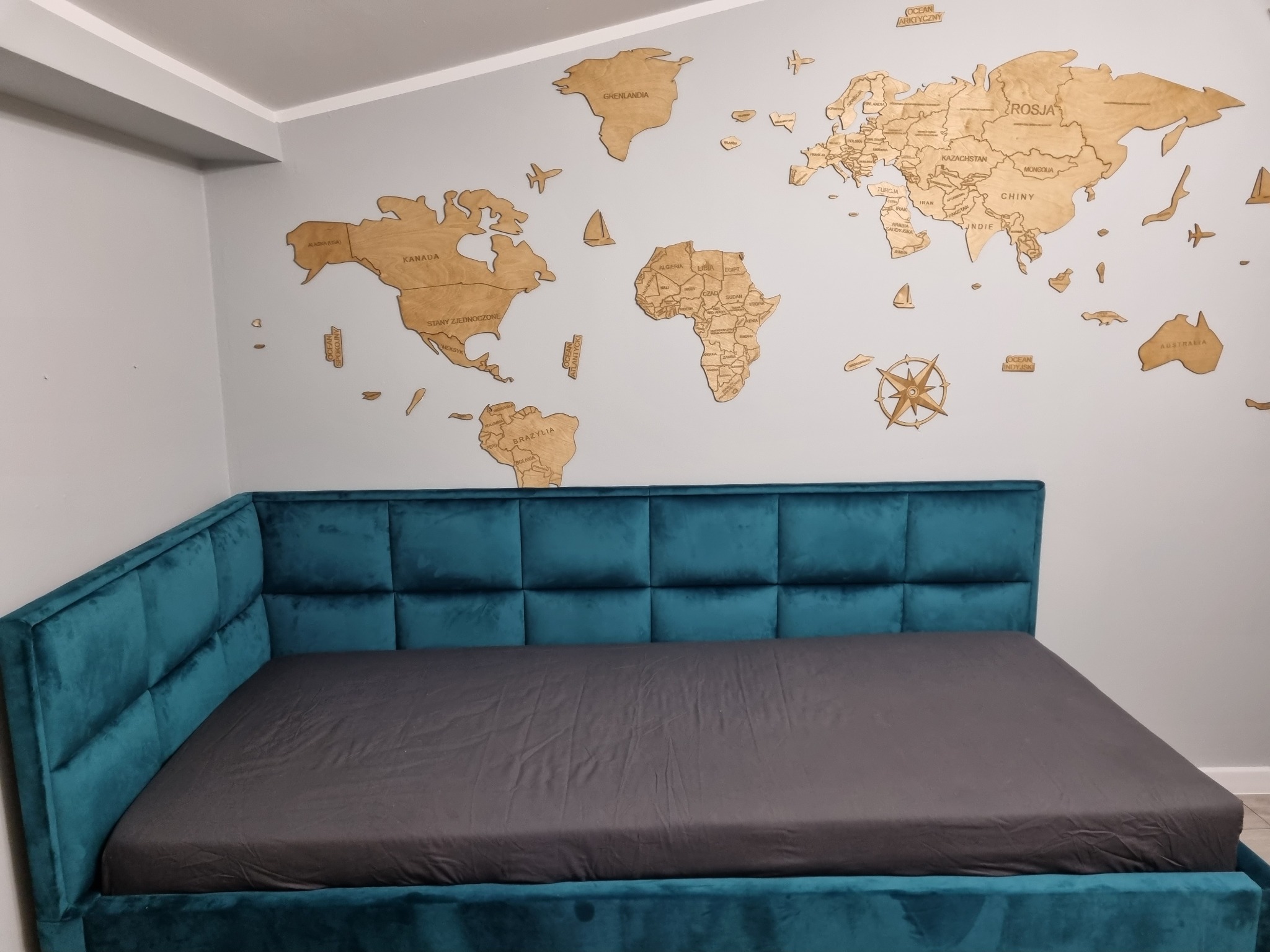 Łóżko tapicerowane w kolorze turkusowym z wezgłowiem w kształcie kwadratów, na ścianie drewniana mapa świata z nazwami państw i oceanów.