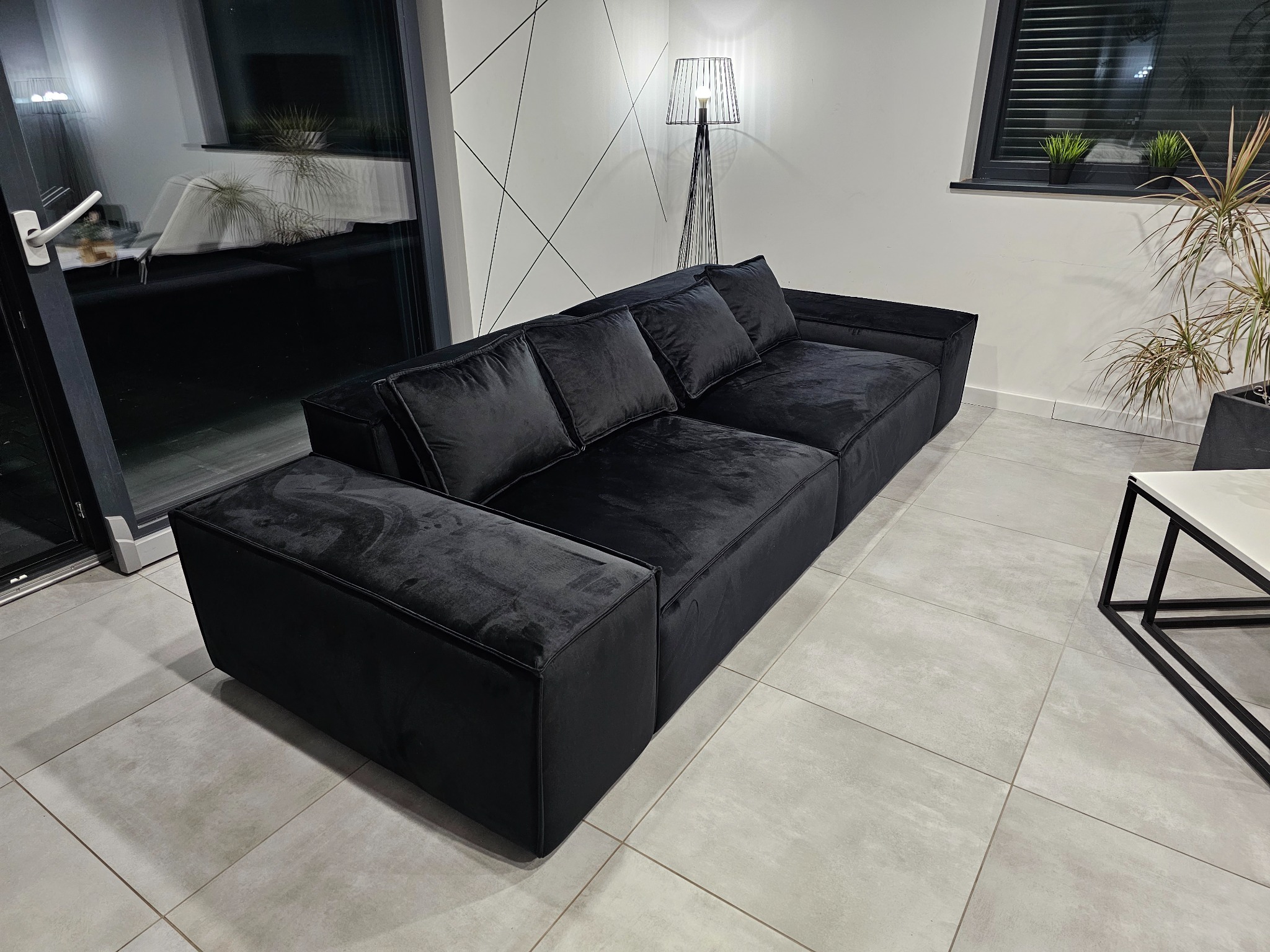 Czarna, modułowa sofa z welurową tapicerką, uzupełniona pufem, na tle białej ściany z geometrycznym wzorem i nowoczesnej lampy podłogowej.