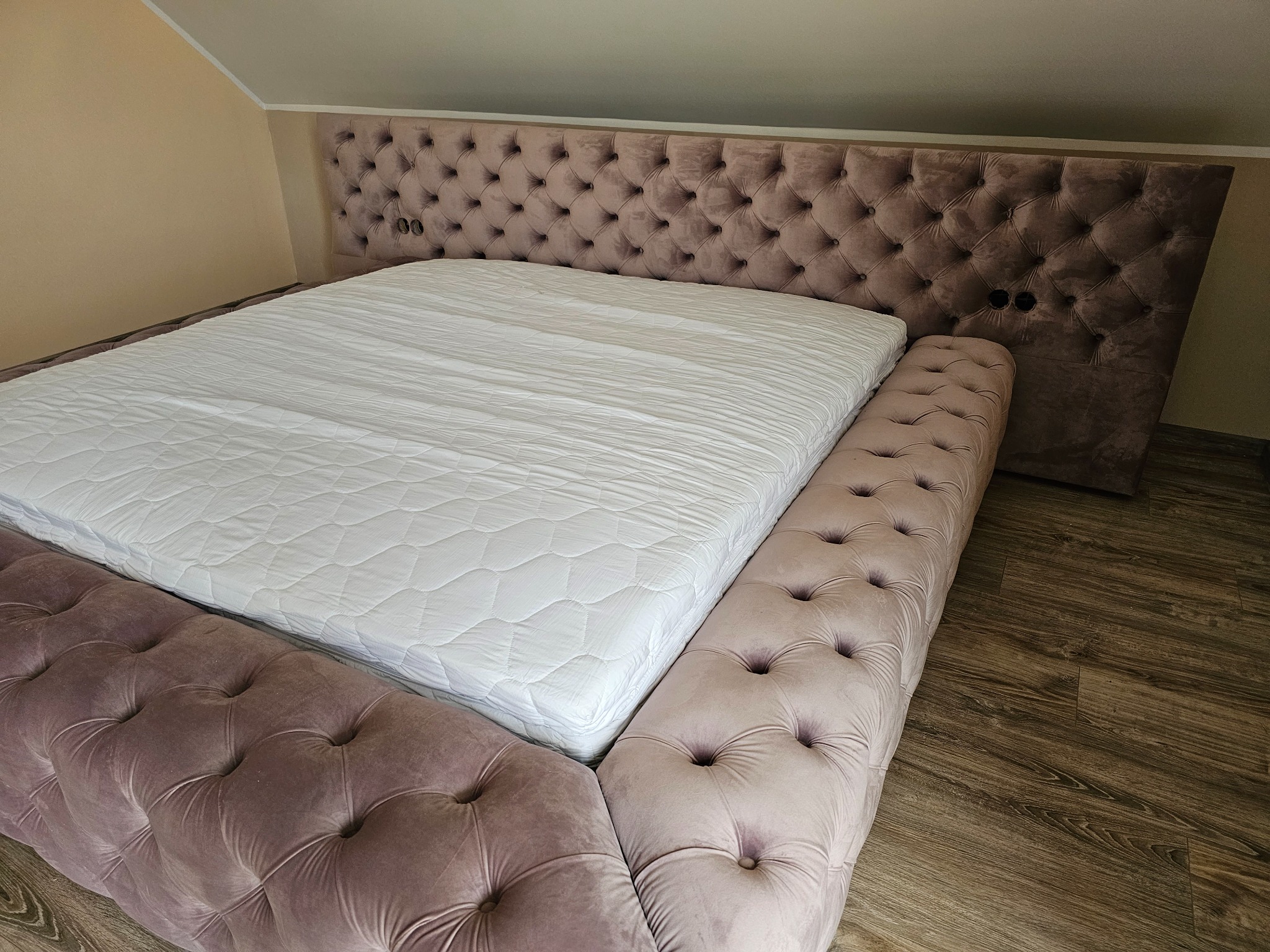 Łóżko z tapicerowanym, pikowanym zagłówkiem i ramą w odcieniach pudrowego różu, z materacem w białej narzucie, zrealizowane w Unisławiu.