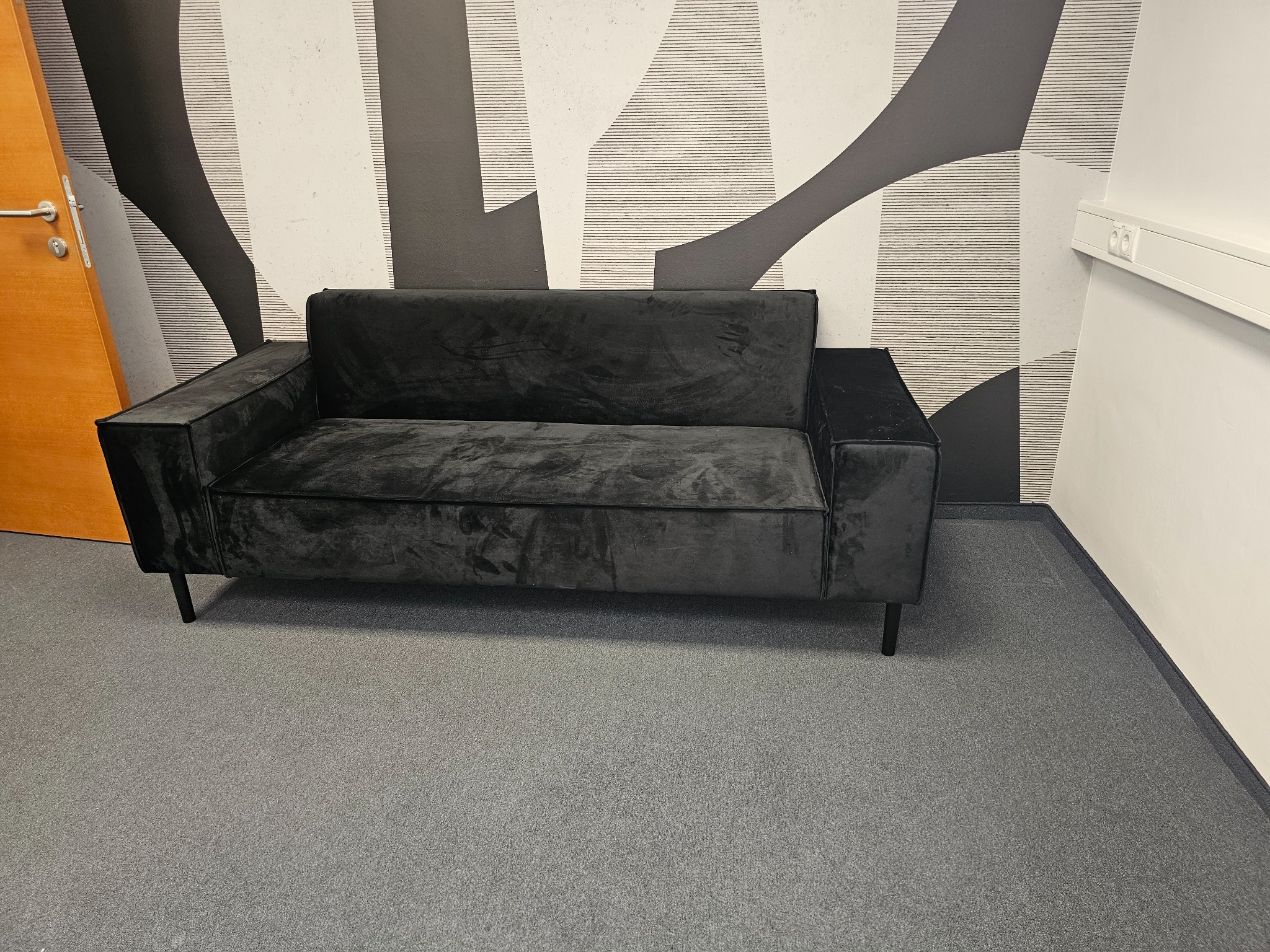 Czarna, welurowa sofa o geometrycznych kształtach stoi na szarym dywanie na tle ściany z abstrakcyjnym, czarno-białym wzorem i fragmentu drewnianych drzwi.