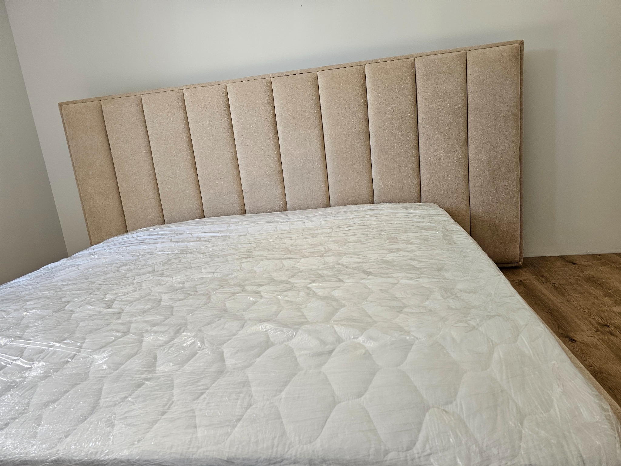 Beżowe tapicerowane wezgłowie łóżka z pionowymi panelami, materac zabezpieczony folią ochronną, podłoga drewniana.