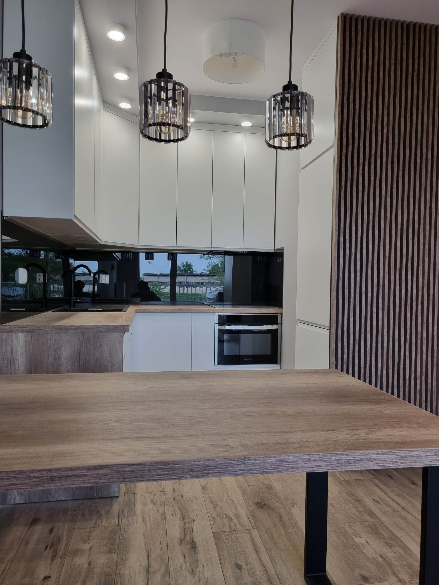 Nowoczesna kuchnia z białymi szafkami, czarnym panelem nad blatem i drewnianym blatem. Nad blatem wiszą trzy ozdobne lampy ze szklanymi elementami. Ściana po prawej stronie zdobiona drewnianymi...
