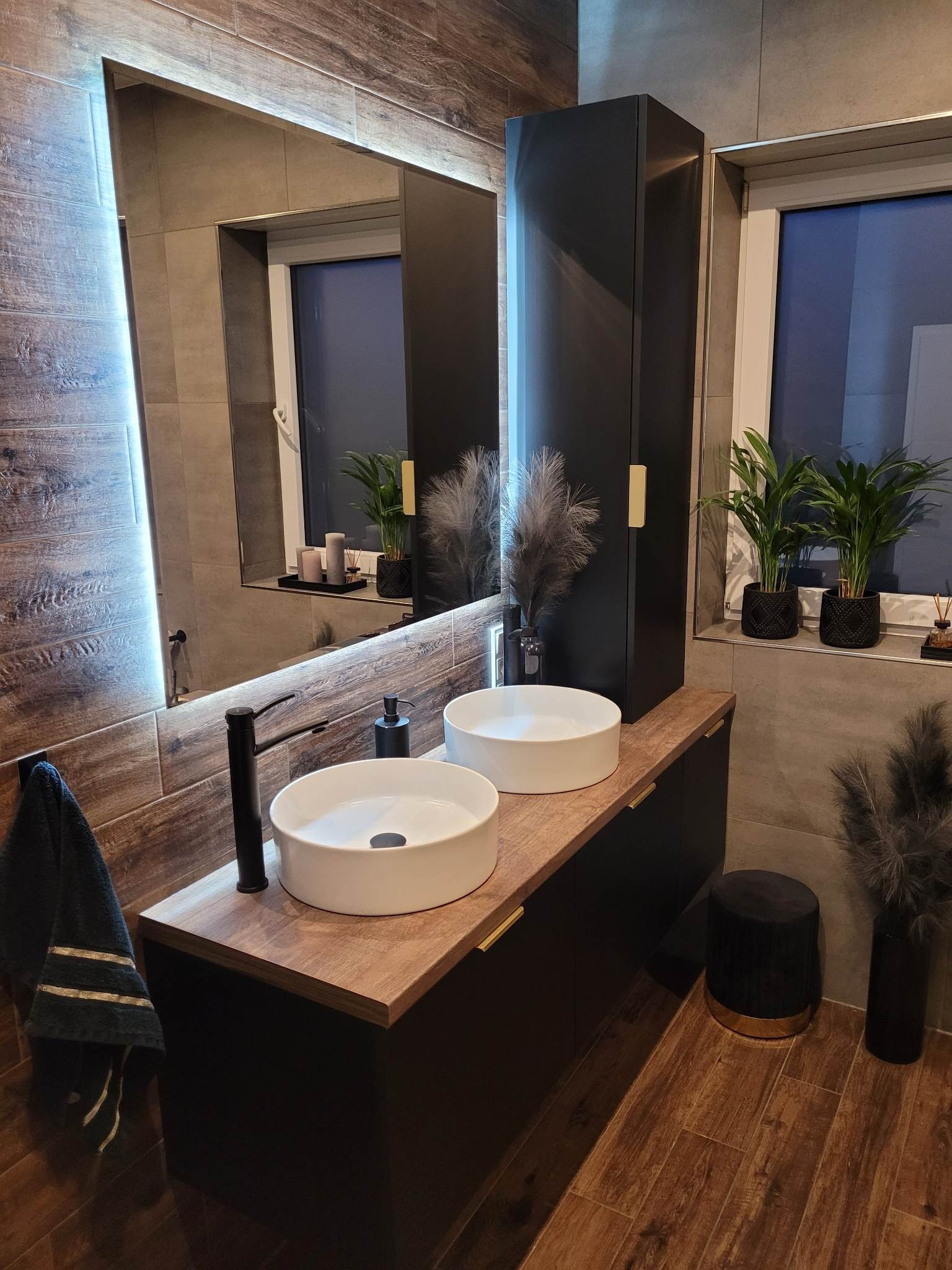 Nowoczesna łazienka z dwoma okrągłymi umywalkami nablatowymi, czarną armaturą i szafką z drewnianym blatem, uzupełniona oświetlonym lustrem i elementami dekoracyjnymi, takimi jak rośliny doniczkowe...