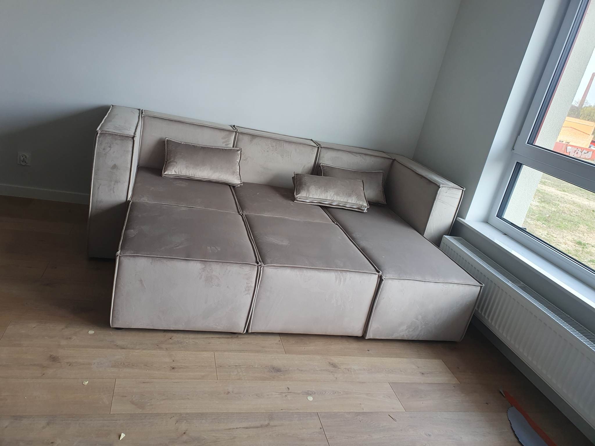 Rozłożona, modułowa sofa w odcieniach beżu z kwadratowymi segmentami i poduszkami, ustawiona na drewnianej podłodze w jasnym wnętrzu z dużym oknem.