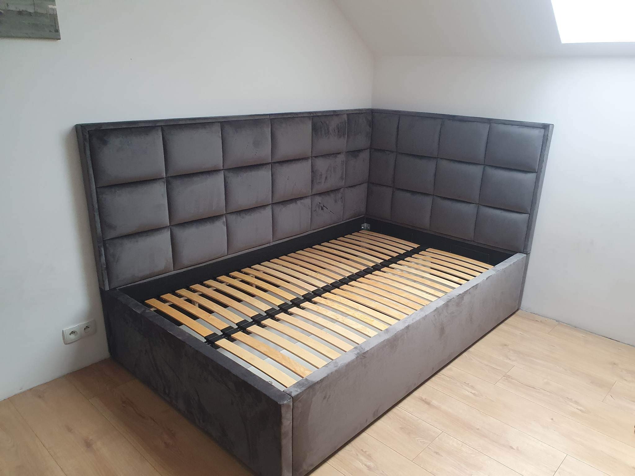 Szare, tapicerowane łóżko z zagłówkiem w kształcie litery L, z drewnianym stelażem, ustawione w rogu pokoju ze skośnym sufitem.