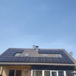 Dom jednorodzinny z zamontowanymi panelami słonecznymi na dachu z dachówką ceramiczną, widoczne okna dachowe, komin, antena satelitarna i drabina oparta o ścianę.