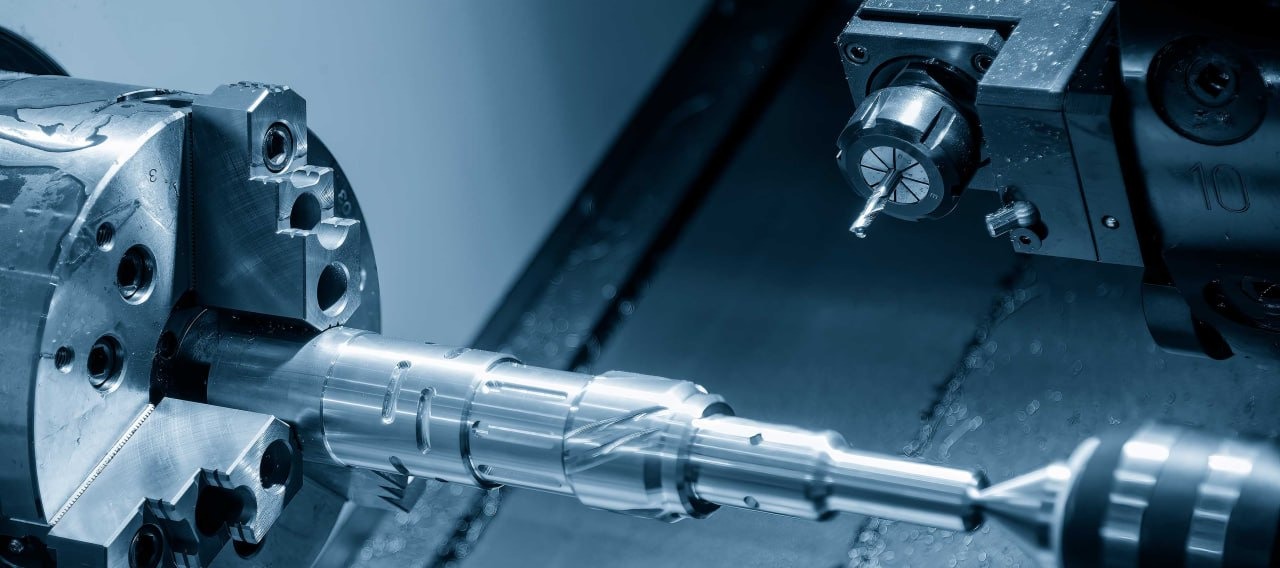 Obróbka metalowego wałka na tokarce CNC; widoczne narzędzie skrawające, uchwyt tokarski i obrabiany detal z rowkami.