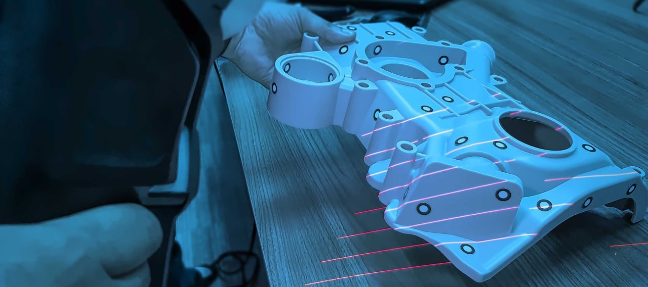 Biały element obudowy silnika z otworami, skanowany laserowo na drewnianym blacie, trzymany w ręce.
