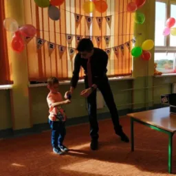 Mężczyzna w czarnym garniturze, prawdopodobnie iluzjonista, wykonuje trik dla chłopca na tle sali udekorowanej balonami i napisem 'Dzień Olaka'.