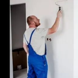 Mężczyzna w niebieskim kombinezonie maluje ścianę wałkiem, widoczne puszki instalacyjne w ścianie, drzwi w tle.